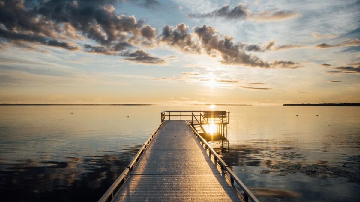 A wooden pier stretching out over calm water toward a soft sunrise under a partly cloudy sky