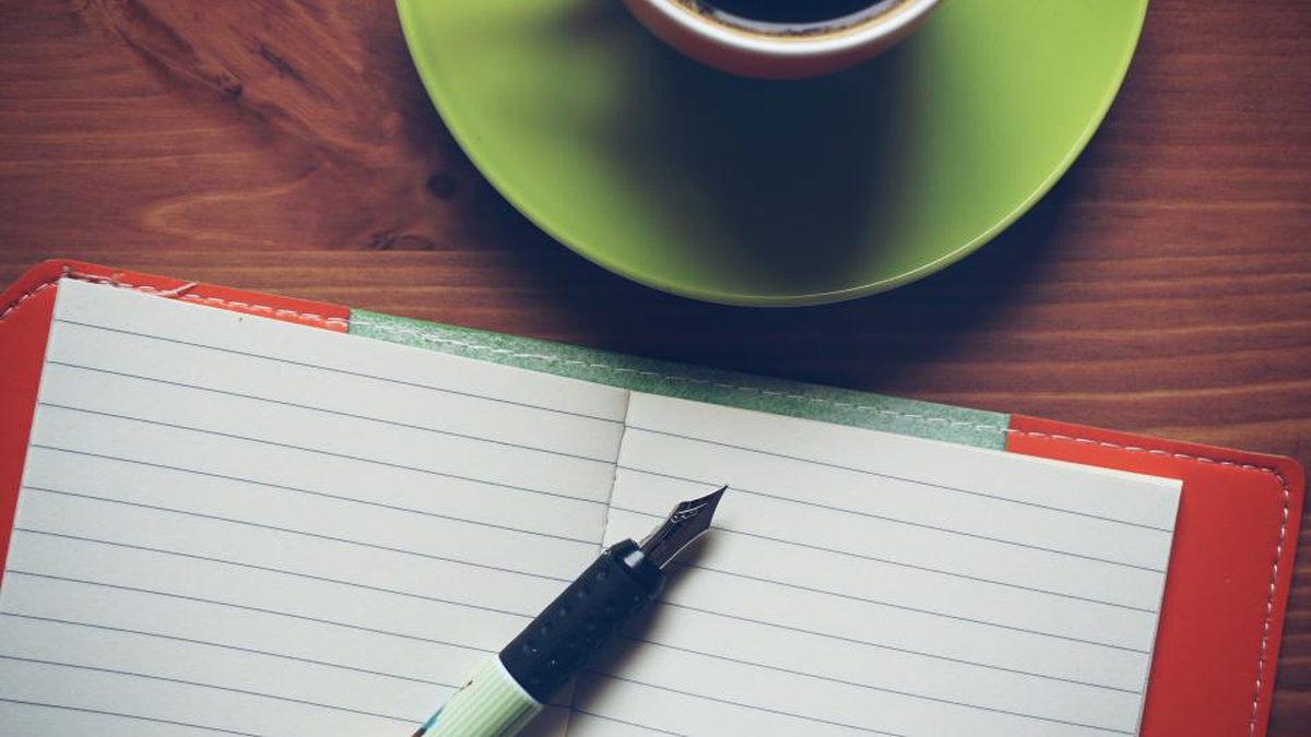 An open lined notebook with a fountain pen beside a green coffee cup and saucer on a wooden table